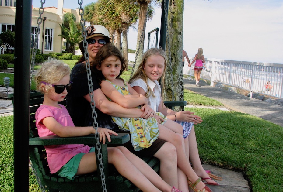 woman with three young girls sits on swing near beach